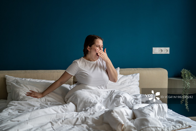 昏昏欲睡的胖女人坐在床上，打呵欠，用手捂住嘴，准备在家度过慵懒的周末图片素材