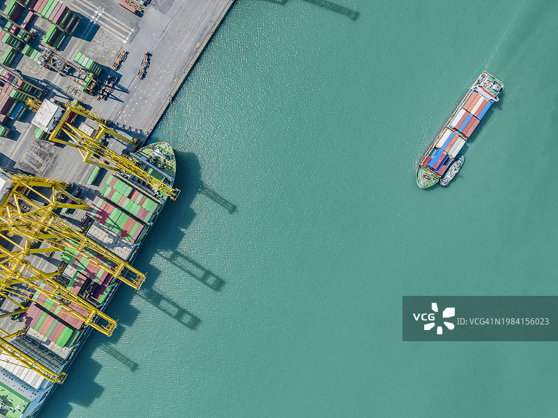 拖船将小型集装箱船推入港口航拍，起重机在码头仓库装载集装箱图片素材