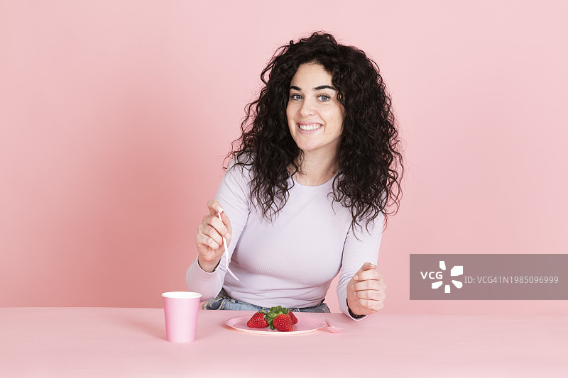 在桌旁手持草莓和咖啡杯的微笑年轻女人图片素材