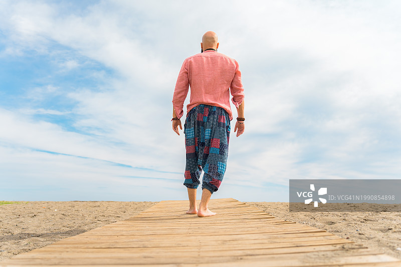 一个秃顶嬉皮士走在沙滩上的木板路上，走向大海图片素材