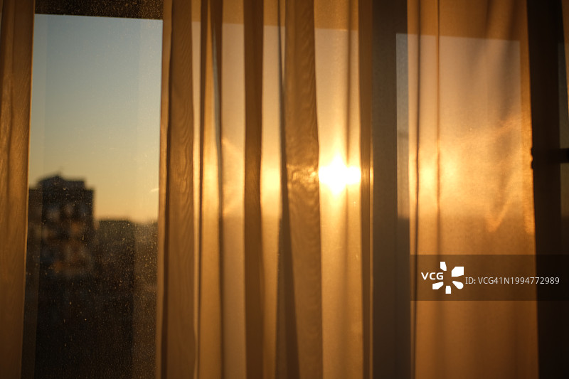 城市公寓窗户中夕阳穿过薄纱窗帘的温暖光芒，展现宁静的日落氛围图片素材