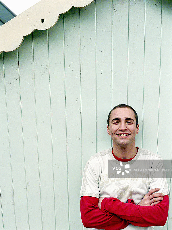靠在海滩小屋，手臂交叉，微笑的年轻男人肖像图片素材
