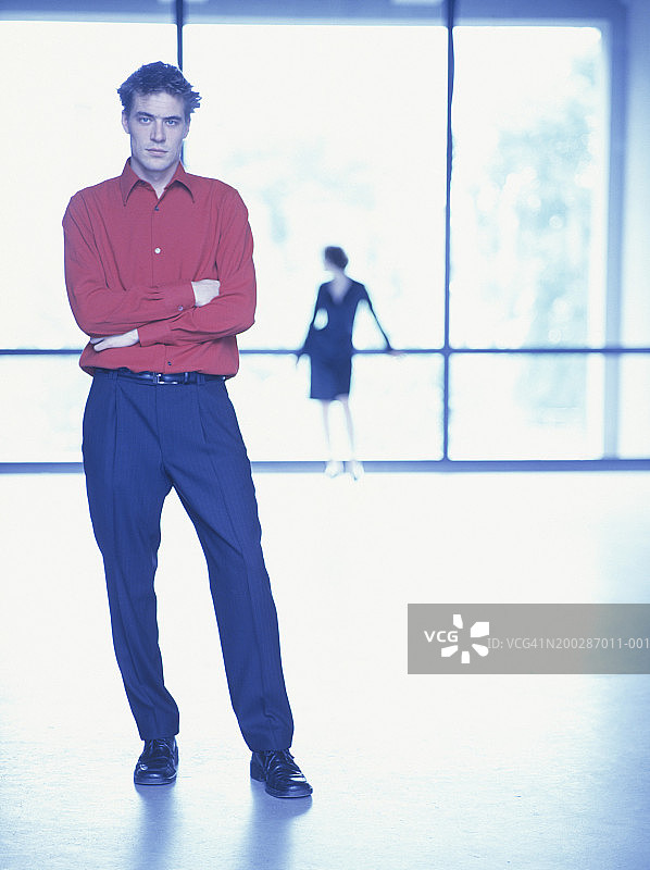 室内交叉双臂站立的男人，背景中的女人图片素材