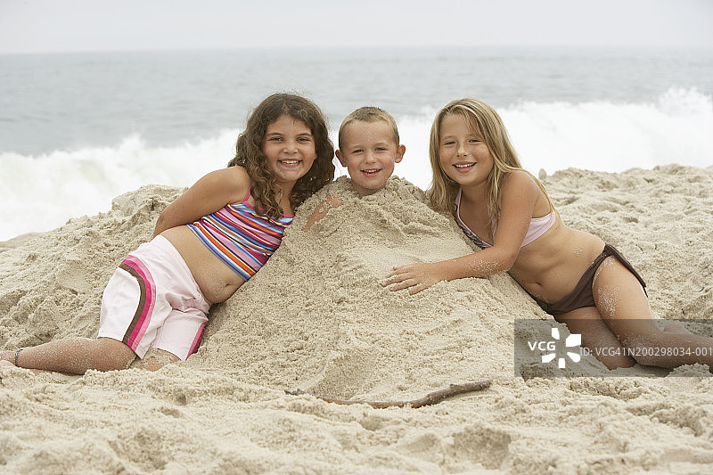 两个女孩(7-9岁)在被沙子掩埋的男孩(2-4岁)的两侧，肖像图片素材
