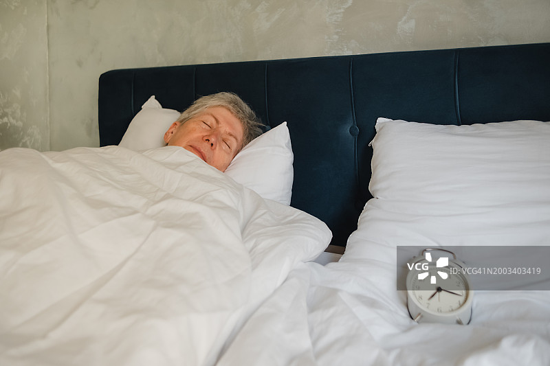 在闹钟旁边的床上，一位正在睡觉的年长女性肖像图片素材