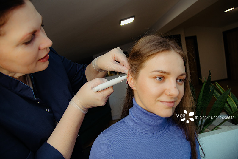 戴手套的师傅保持冷静，用穿孔枪在微笑的顾客的软骨上打孔图片素材