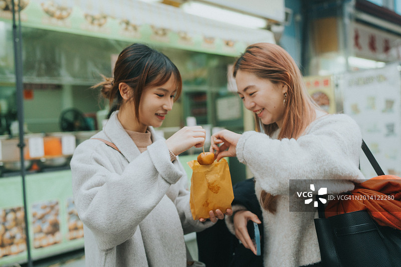 享受休闲小吃：中国香港的鱼蛋，两位亚洲年轻女性站在小吃摊前图片素材