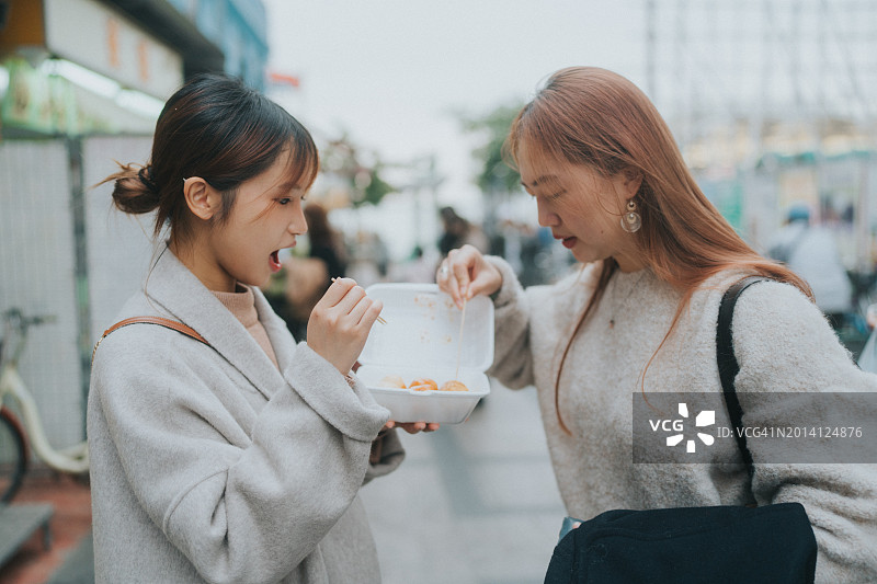两名亚洲年轻女性在户外分享美食，品尝香港街头小吃鱼丸图片素材