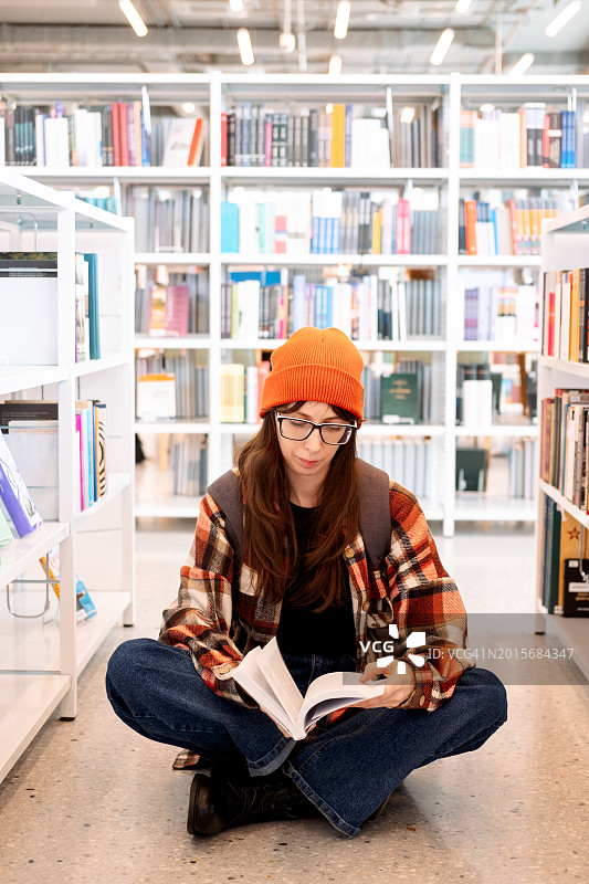 戴红色帽子、穿格子衬衫的年轻女子坐在图书馆的地板上读书图片素材