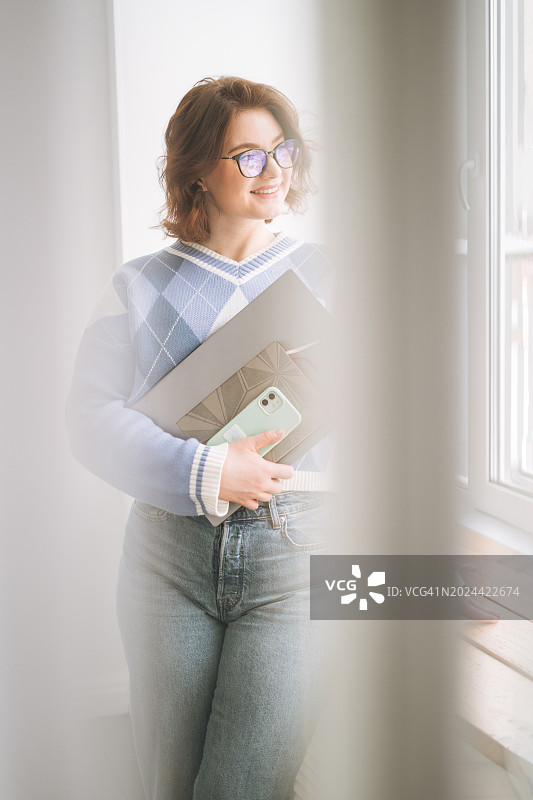 戴眼镜的微笑年轻女学生在窗边使用笔记本电脑图片素材