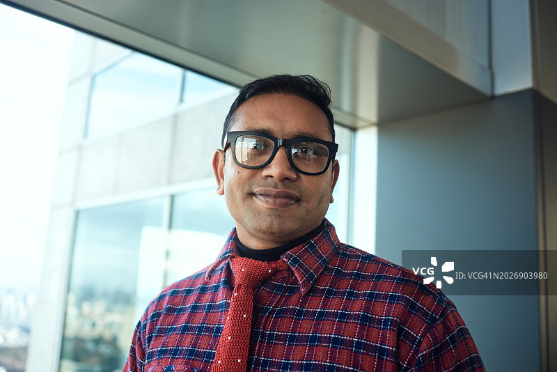 一位印度男子在高层办公室的窗户旁交叉双臂的肖像图片素材