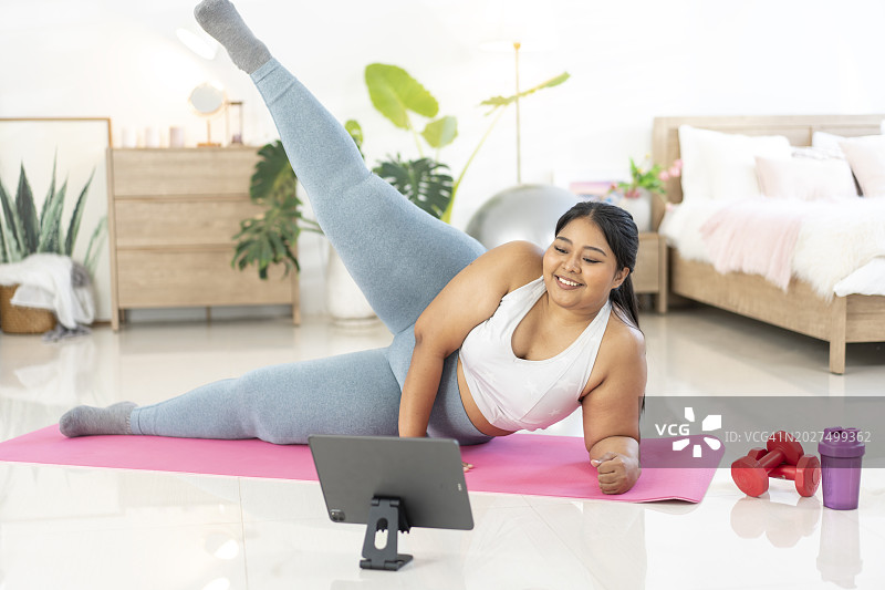 有魅力的超重女人在家中，面前放着笔记本电脑，根据视频在垫子上锻炼，抬起腿图片素材