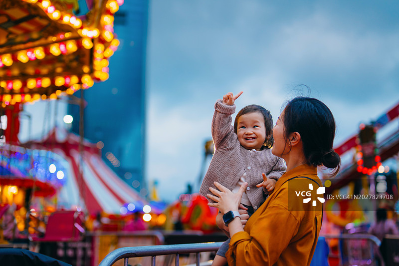 快乐的年轻亚洲母亲抱着她可爱的女婴，她们在嘉年华玩得很开心，一起享受时光。家庭生活方式。家庭欢乐时光。创造难忘的时刻图片素材