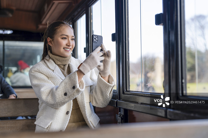 快乐的亚洲女性独自坐在穿梭列车上，欣赏花园公园的景色，在韩国济州岛旅行时使用智能手机拍照图片素材