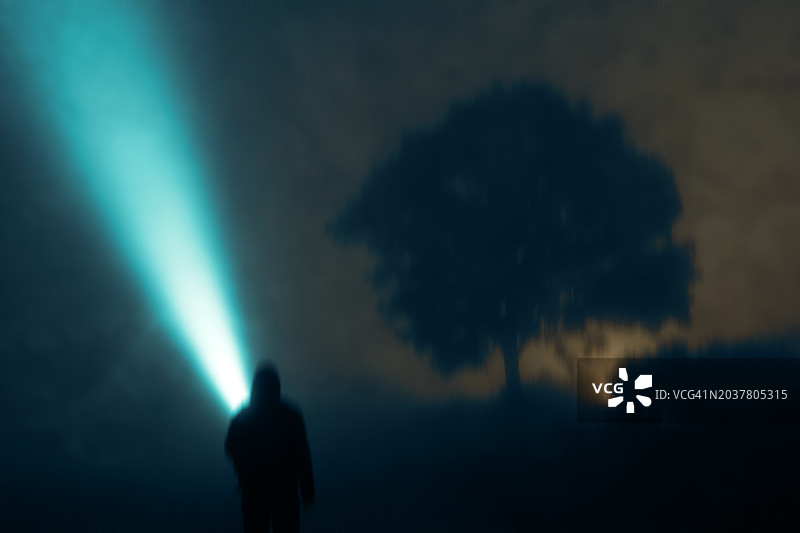 一个神秘的孤独蒙面人，用手电筒光束指向天空。在乡村一个怪异多雾的夜晚。带有粗糙模糊的编辑效果图片素材