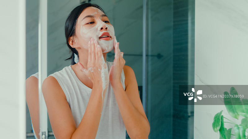 年轻的亚洲女人在家里早上在盥洗室的镜子前享受面部泡沫清洁，家里的日常生活图片素材