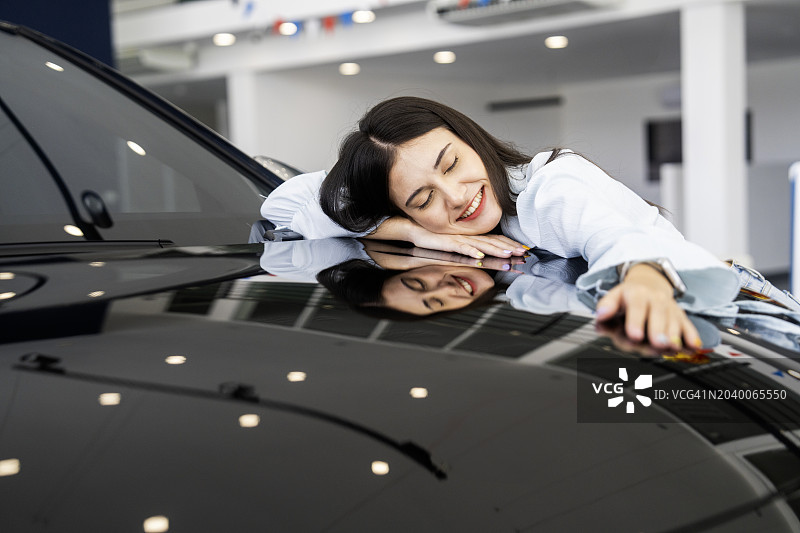 在汽车经销店拥抱汽车并表达爱意的漂亮女人。快乐的女性在展厅选择她梦想中的汽车购买。图片素材