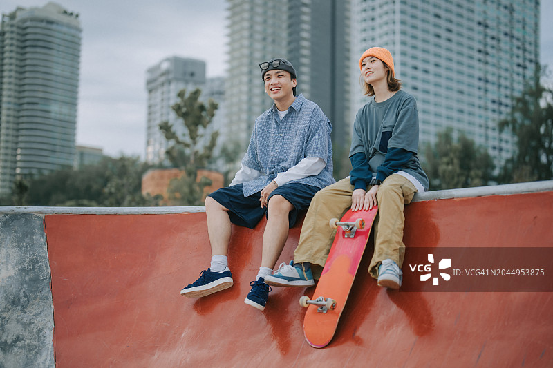 在滑板公园并排坐在墙上休息的亚洲滑板情侣图片素材