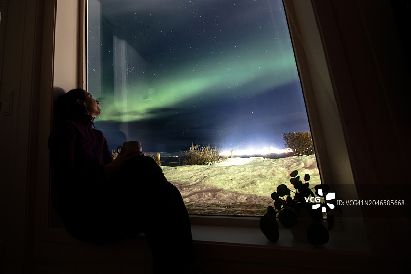年轻女子在家中从窗户向外看北极光图片素材