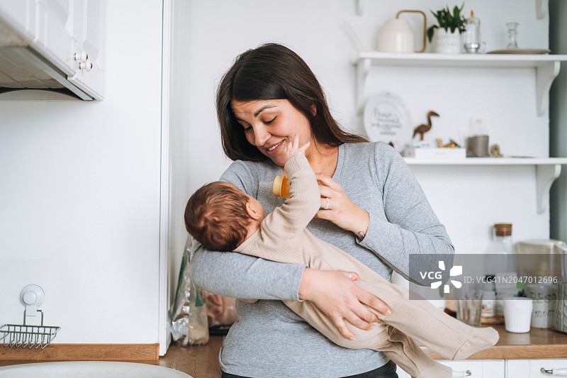 年轻的棕发母亲在家中厨房里用奶瓶给3-4个月大的婴儿喂水图片素材