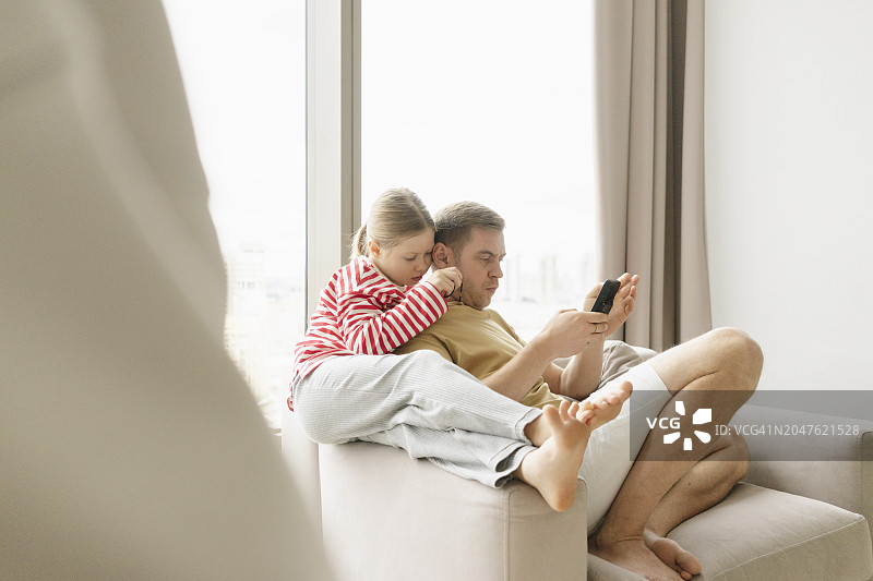 女孩坐在父亲旁边，父亲在家使用智能手机图片素材