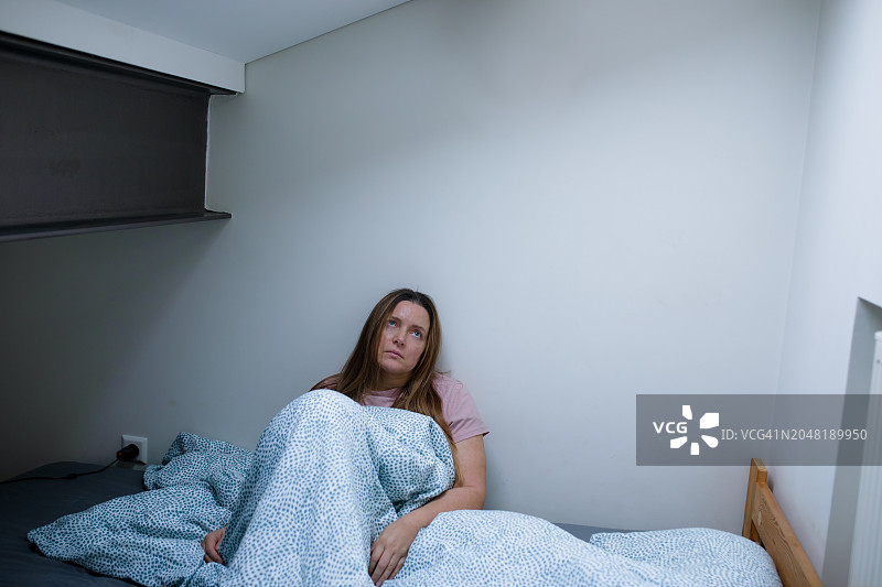 情绪低落的女人裹着毯子坐在床上，眼睛向上图片素材