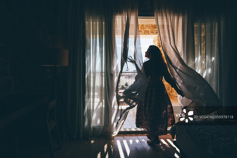 女子打开窗帘向外看，在日出时欣赏酒店房间的阳台图片素材