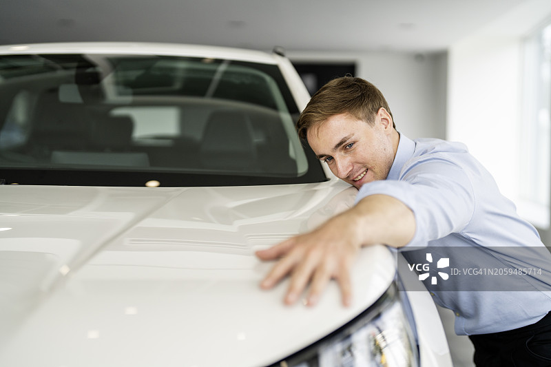 快乐的年轻男子拥抱新车引擎盖，梦想在汽车展厅购买车辆图片素材