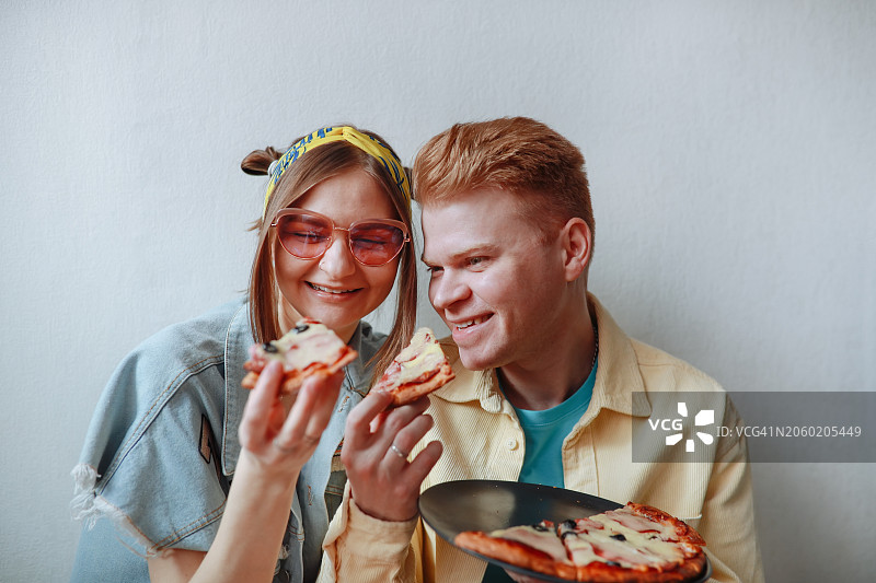 时尚Z世代情侣在地板上靠墙而坐一起享用披萨，穿着色彩鲜艳的服装和粉色太阳镜的休闲零食图片素材
