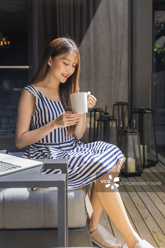 休闲放松的亚洲女人自由职业者使用笔记本电脑和智能手机咖啡杯，躺在游泳池附近的木制甲板椅上，美丽的家庭建筑设计与花园庭院图片素材