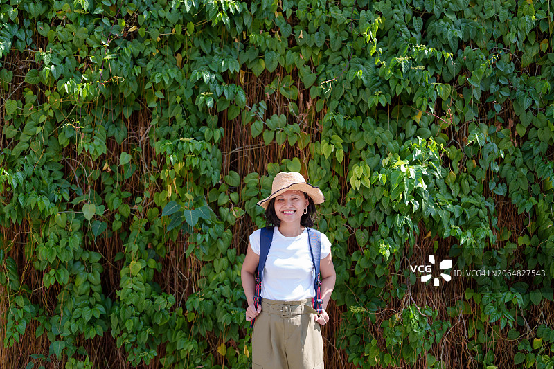 一位快乐的亚洲女性独自旅行者，戴着草帽和背包，在槟城乔治市的绿叶墙前摆姿势图片素材