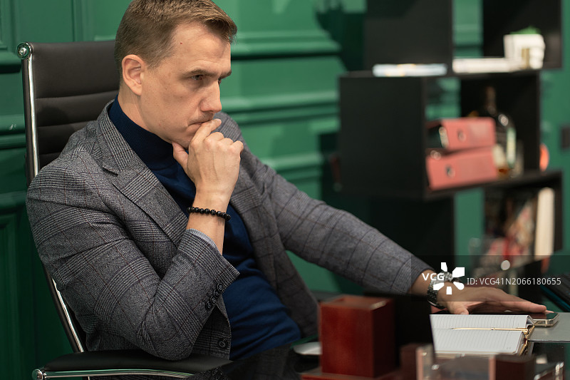 穿夹克和蓝色高领衫的男人在办公室的电脑桌前思考图片素材