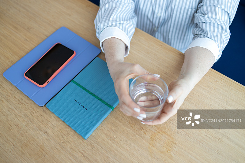 女人在办公桌旁手持一杯水，周围有办公用品和智能手机图片素材
