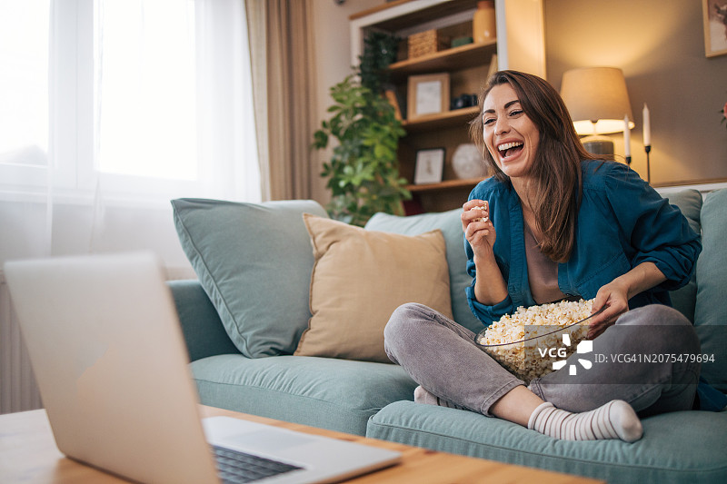 坐在家里的沙发上看喜剧电影，吃着爆米花微笑的女人图片素材