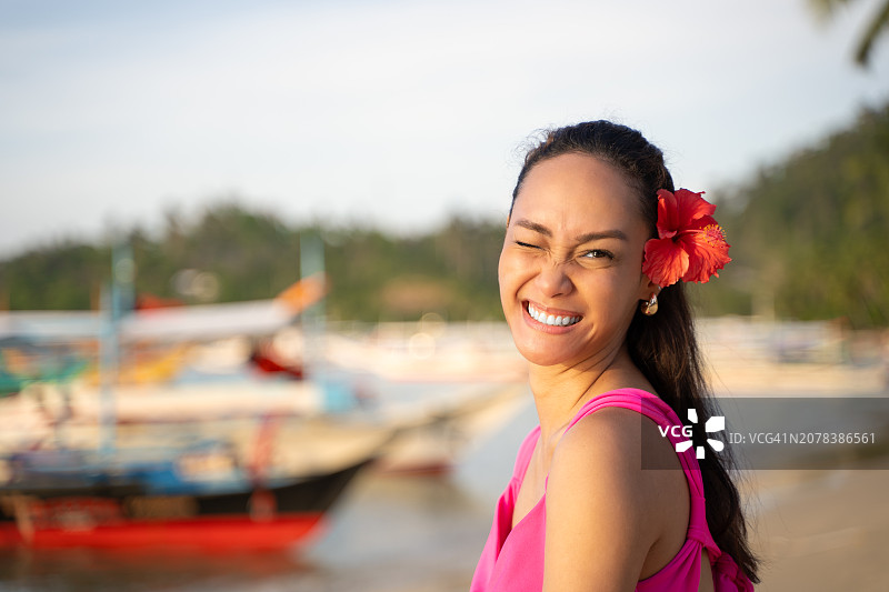 在海滩上微笑、眨眼，头发上戴着鲜花，看着镜头的美丽菲律宾女人图片素材