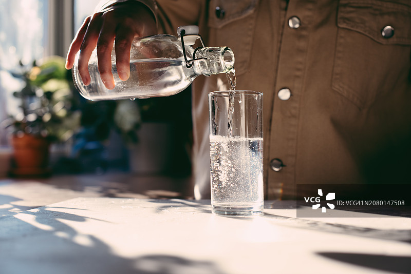 女性在家中将新鲜水从水壶倒入玻璃杯的特写。有魅力的女孩在家里厨房喝水。年轻女性在厨房里手持柠檬水慢动作图片素材
