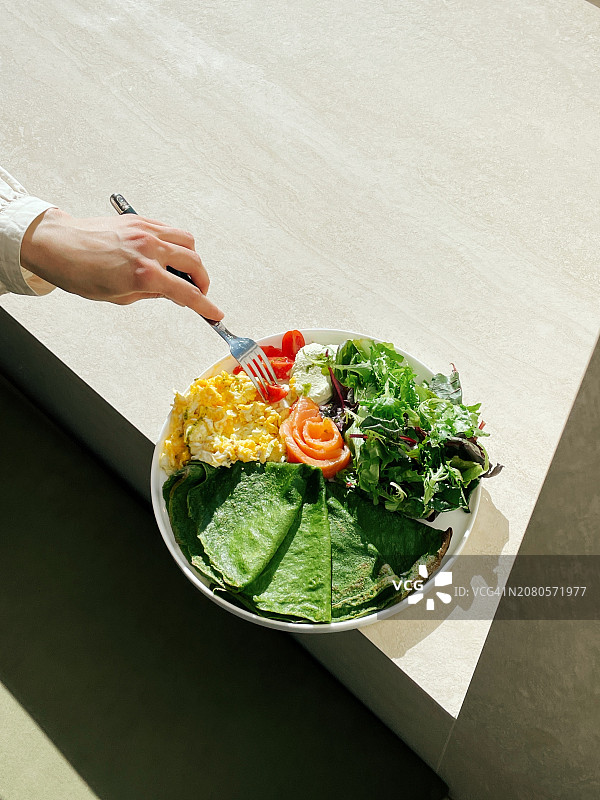 女人吃菠菜煎饼、炒鸡蛋、三文鱼、番茄和生菜的鸟瞰图图片素材