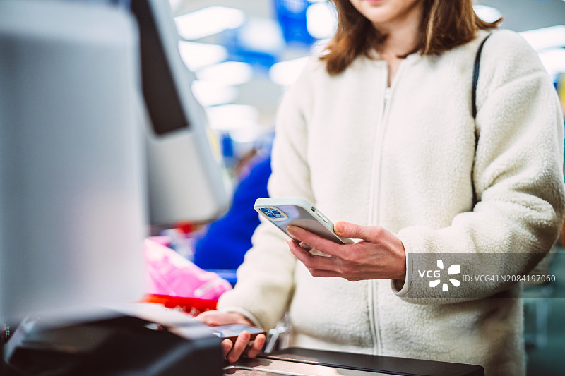 一位女顾客在超市自助结账时使用智能手机进行非接触式支付图片素材
