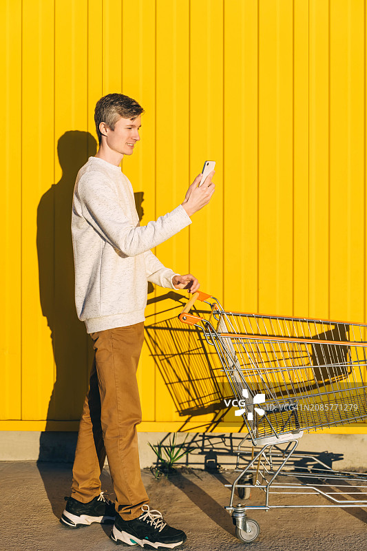 在黄色背景下手拿手机和购物车里的年轻男人，购物概念图片素材
