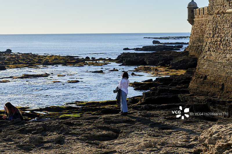 女人在日落时分在多石的海滩上漫步图片素材