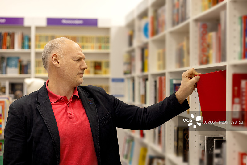 在书店里，一位头发灰白的中年男人从书架上取下一本红色的书图片素材