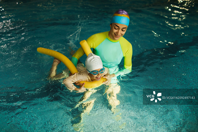 孩子在游泳池里快乐地学习游泳。老师用游泳棒帮助保持平衡。图片素材