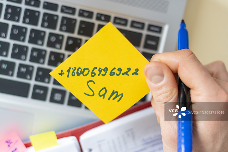 联系方式捕获 - 商务工作人员的手拿着一张明亮的纸条，上面有针对笔记本电脑背景的电话号码图片素材