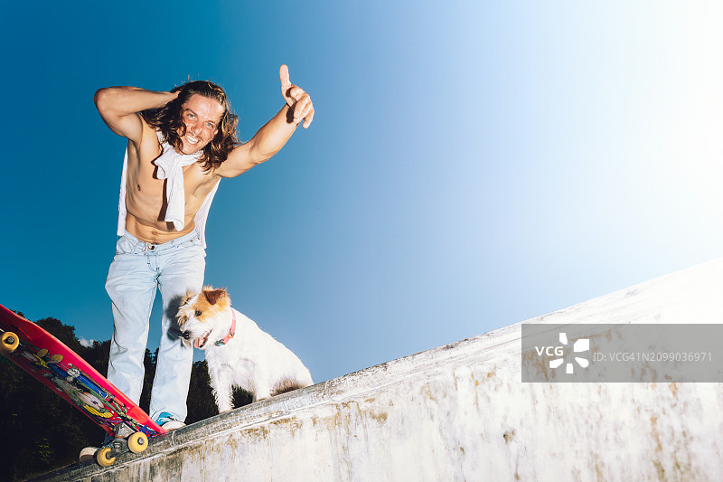 滑板公园生活方式：无上装男子和快乐的狗在晴朗的蓝天下玩滑板图片素材