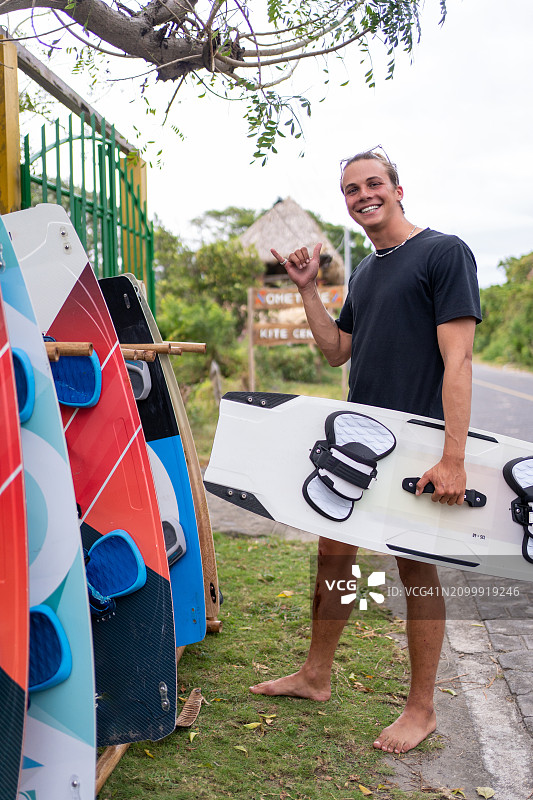 一位年轻的金发男子带着他的冲浪板对着镜头微笑，并做出“放松”的手势图片素材