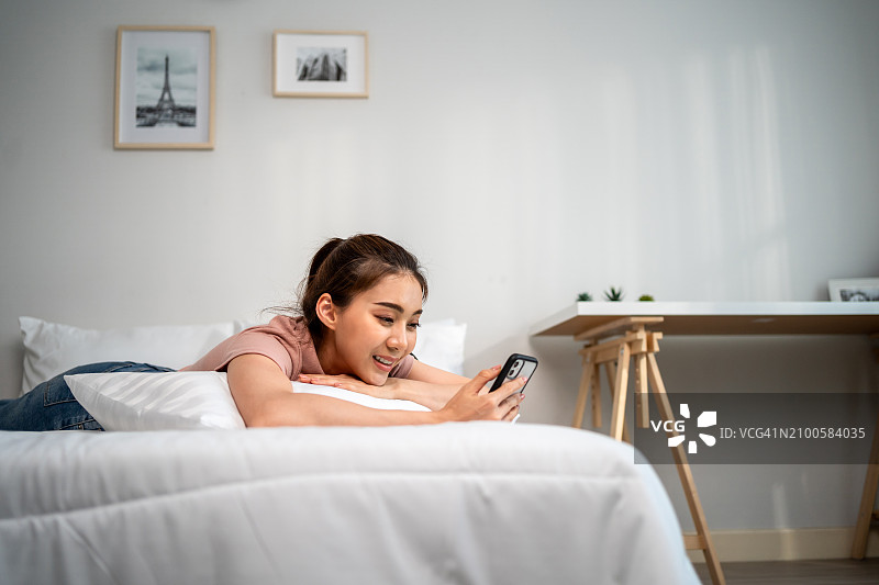 早上在家在床上用手机聊天的迷人亚洲女性。 躺在卧室里享受周末假期的快乐休闲美女，使用智能手机进行交流图片素材