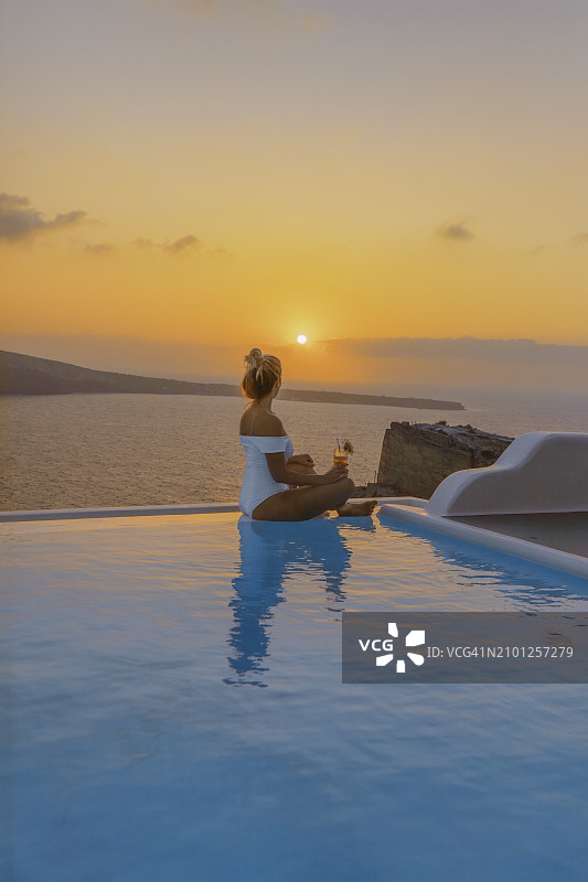 年轻女子坐在无边泳池边，欣赏日落和地中海图片素材