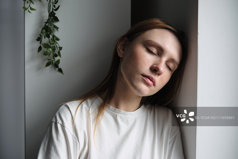 情绪倦怠：站在窗边，闭着眼睛，感到精疲力竭的千禧一代女孩图片素材