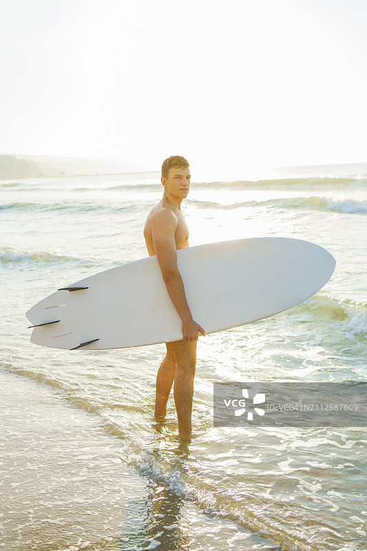 日落时分站在水中的年轻男子与冲浪板图片素材