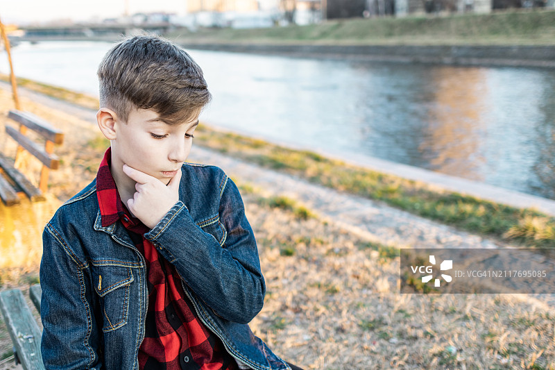 坐在日落时分河边的长椅上，用笔记本电脑与朋友交流的可爱男孩。在线教育。图片素材